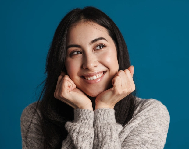 Young woman smiling at camera with hands near face against blue background.