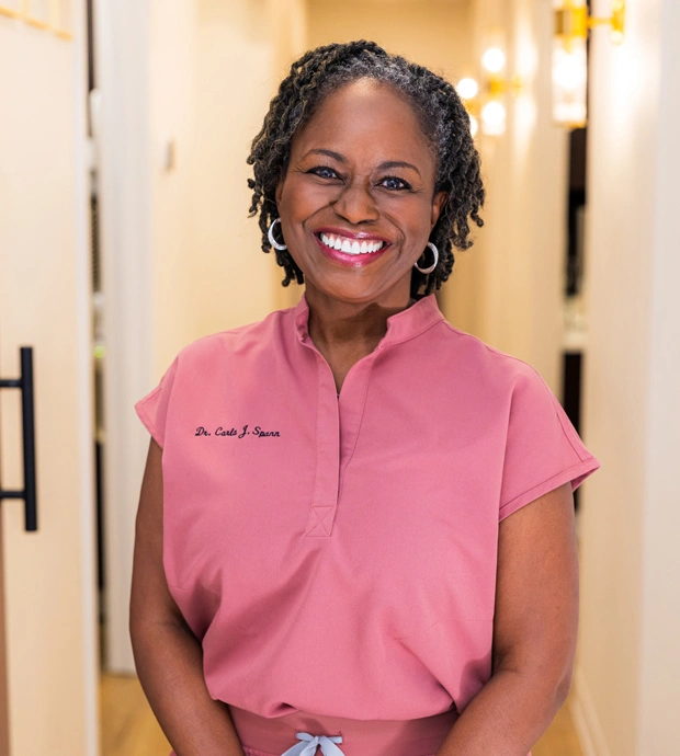 Smiling dental professional in pink scrubs stands in modern dental office hallway.