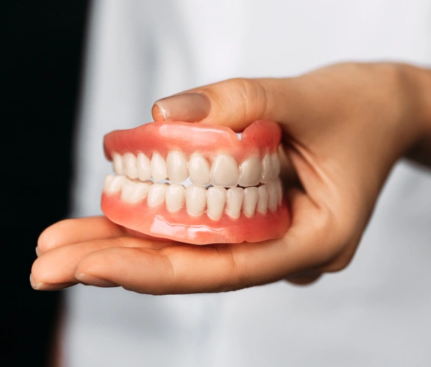 Hand holding complete denture model displaying full set of artificial teeth against white background.