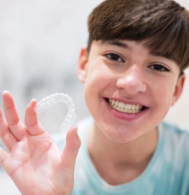 Young boy holding clear dental aligners, smiling at camera in dental office setting.