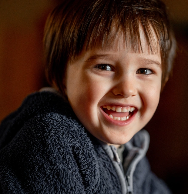 Young child smiling brightly at camera, wearing dark sweater, warm indoor setting.