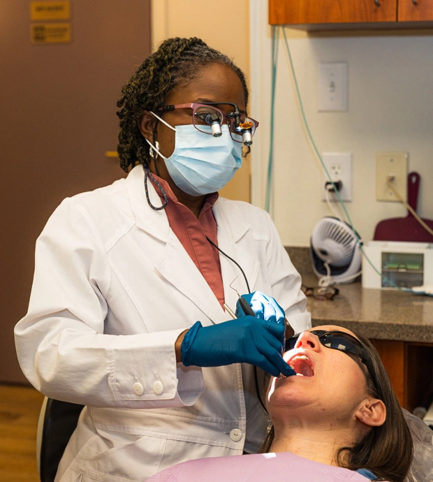 Dental hygienist in white coat and mask examines patient's teeth in clinical office setting.
