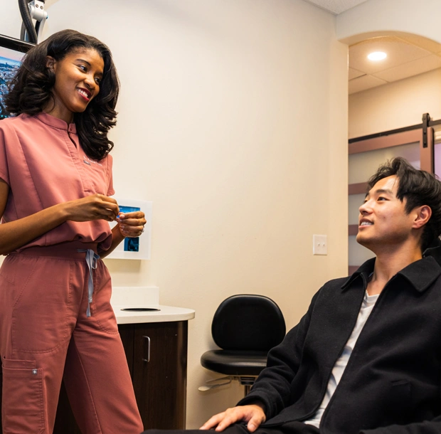 Two professionals in dental office setting; woman standing holding tablet speaks with seated man in business attire.