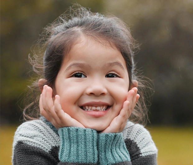 Happy young girl smiling outdoors wearing green sweater, hands on cheeks.