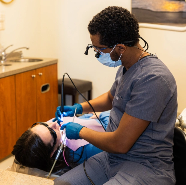 Dentist in blue scrubs and mask examining patient's teeth in dental office treatment room.