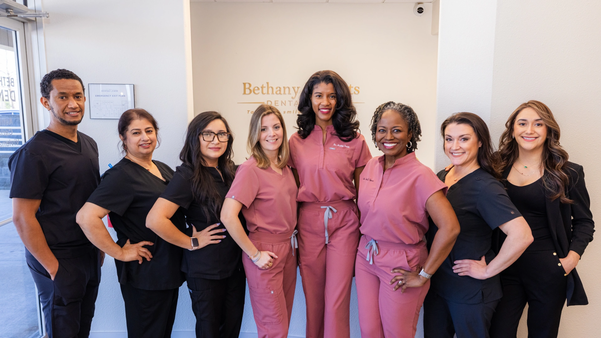 Dental team of eight professionals posing together in modern office setting wearing coordinated scrubs.