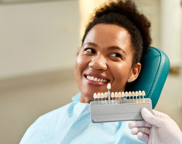 Patient smiling in dental chair holding shade guide for teeth whitening consultation.