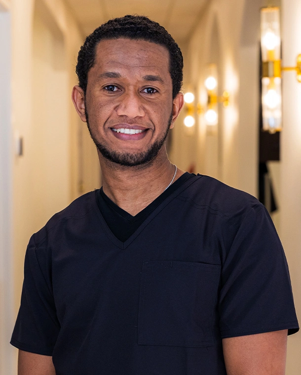 Smiling man in black v-neck shirt poses in modern dental office hallway with warm lighting.