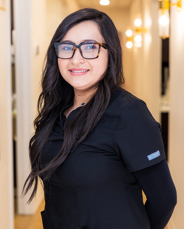 Professional woman wearing glasses and black polo shirt smiling in dental office hallway.