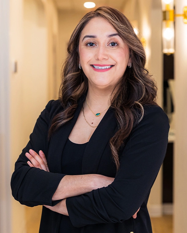 Professional woman in black blazer with arms crossed, smiling in modern dental office hallway.
