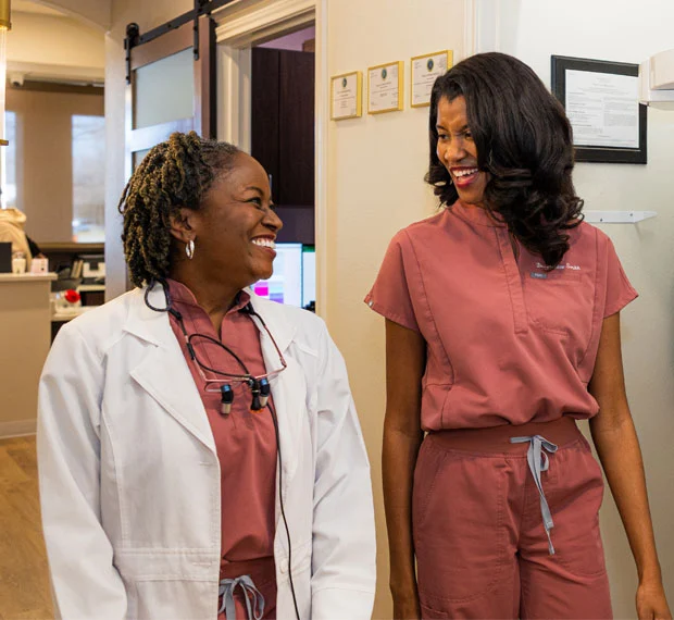 Two women in a dental office; one wearing a white coat and the other in scrubs, conversing professionally.