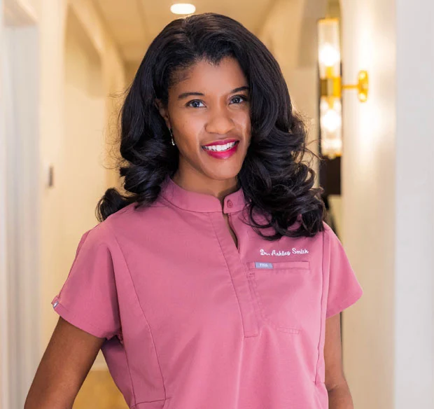Professional woman in pink dental scrubs smiling in modern dental office hallway.