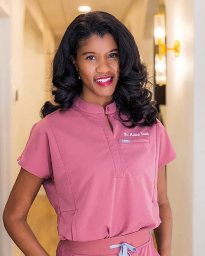 Professional woman in pink dental scrubs smiling in modern dental office hallway.