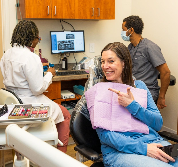 Patient in dental chair smiling while holding pink bib, dental office with staff in background.